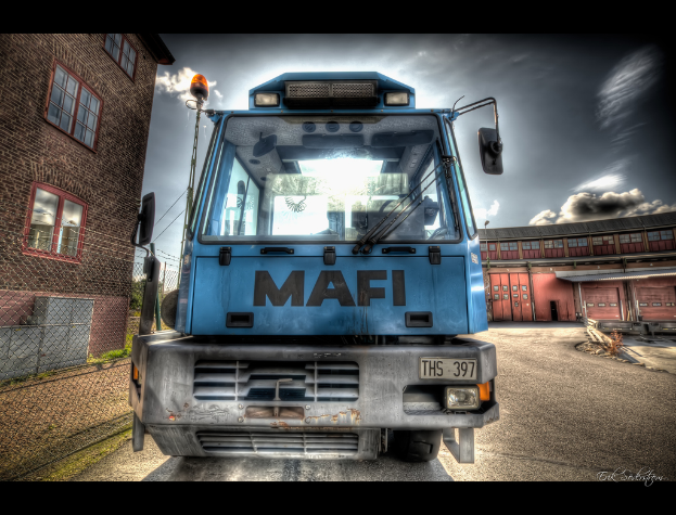 Eine animierte Abbildung eines Busses mit Nummernschild, einem Gebäude mit Fenstern auf der linken Seite, einem Wasserzeichen im rechten Eck und einem Hintergrund mit einem weiteren Gebäude und einem bewölkten Himmel.