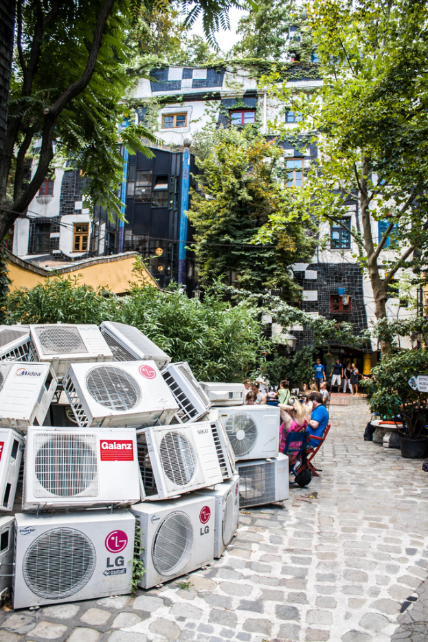 Luftkühler sind links unten auf dem Boden platziert, mit einer Spur rechts daneben, Menschen auf Stühlen neben der Spur sitzen und Gebäude, Bäume und Pflanzen oben.