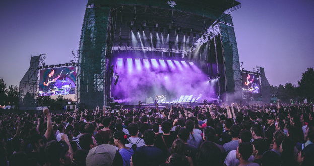 Eine große Menschenmenge bei einem Musikfestival schaut auf eine beleuchtete Bühne mit Instrumenten und Bildschirmen, die vor einem Gebäude, Bäumen und einem klaren blauen Himmel aufgebaut ist.