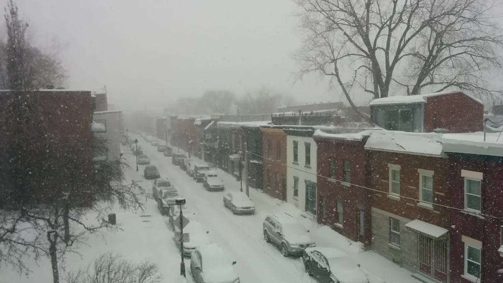 Eine schneebedeckte Straße mit Autos und Gebäuden auf beiden Seiten, mit Bäumen im Hintergrund.