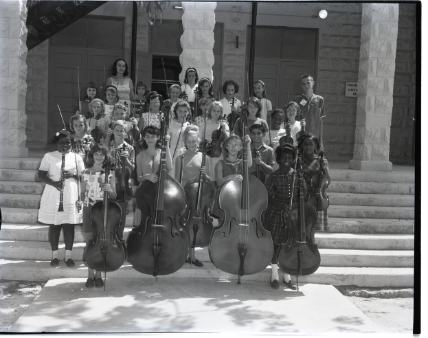 Kinder stehen auf Stufen und halten Musikinstrumente, darunter Celli, Geigen und Trompeten, mit Säulen dahinter und einer Wand mit Türen im Hintergrund.