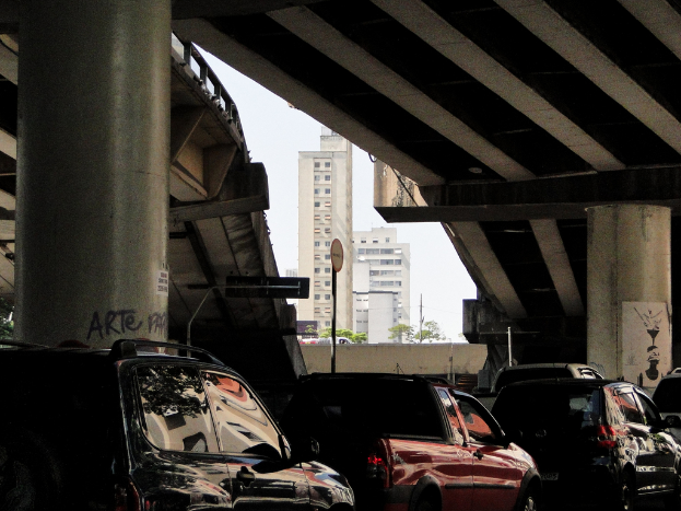 Blick von unten auf eine Überführung mit Säulen, parkenden Autos und einem zentralen Gebäude.
