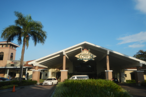 Einfahrt zu einem Resort mit Palmen vorne, umgeben von Gebäuden, Fahrzeugen auf der Straße, Verkehrshütchen, Büschen, Bäumen und einem bewölkten Himmel im Hintergrund.