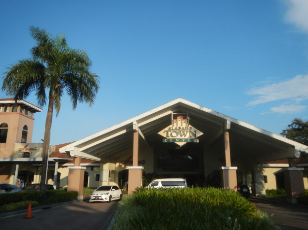 Einfahrt zu einem Resort mit Palmen vorne, umgeben von Gebäuden, Fahrzeugen auf der Straße, Verkehrshütchen, Büschen, Bäumen und einem bewölkten Himmel im Hintergrund.