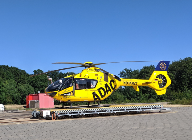 Ein gelber Hubschrauber steht auf dem Rollfeld eines Flughafens, umgeben von Bäumen und einem klaren blauen Himmel, mit verschiedenen Gegenständen, die mit dem Hubschrauber in Zusammenhang stehen, auf dem Rollfeld verstreut.