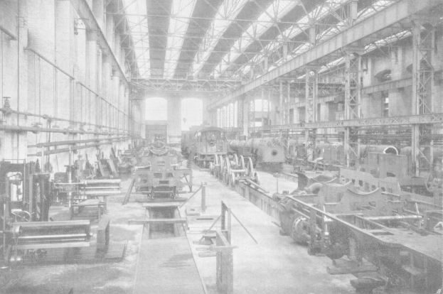 Schwarz-weiß-Foto einer großen Fabrikhalle mit zahlreichen Zügen auf dem Boden, industrieller Maschinen, Stützpfosten, Wänden und einem sichtbaren Dach.