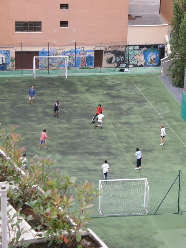 Kinder spielen Fußball auf einem Feld mit Torpfosten, ein Gebäude mit Graffiti im Hintergrund, Pflanzen links und Bäume rechts.