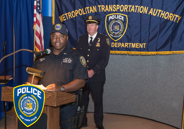 Zwei Polizeibeamte stehen vor einem Podium, einer hält Papiere und spricht in ein Mikrofon, mit einer Fahne, einem Tisch und einer Tafel mit Symbolen und Text im Hintergrund.