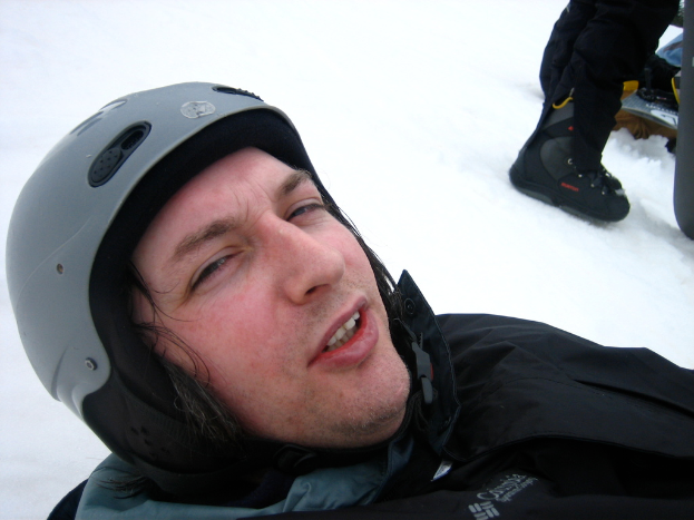 Ein Mann in einem Pullover und Helm steht im Schnee, mit den Beinen und Skiern einer anderen Person in der oberen rechten Ecke.