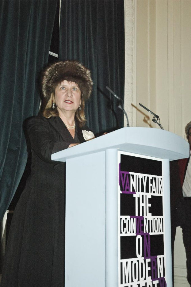 Eine Person steht an einem Podium mit Text darauf und spricht in ein Mikrofon, umgeben von mehreren Menschen und Vorhängen.