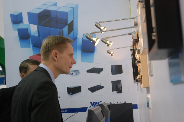 Ein Mann in einem blauen Hemd und schwarzem Blazer steht in der linken Ecke und schaut auf ein Messgerät an einer weißen Wand mit Lampen darüber und einem weißen Banner mit einem Würfel und Monitor dahinter.