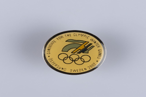 Ein schwarzes Kreisabzeichen mit einem goldenen Olympic Winter Games-Logo und weißer Schrift auf einer weißen Oberfläche.