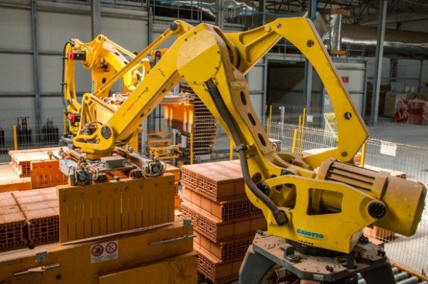 Ein großes Lagerhaus voller numerous boxes, Kränen und diverser industrieller Ausrüstung, mit einem Zaun und Pfählen im Vordergrund und einer Wand im Hintergrund.