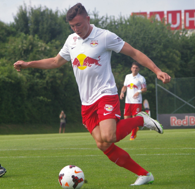 Ein Mann in einem Fußballtrikot kickt einen Ball auf einem grünen Feld mit Bäumen und einem klaren blauen Himmel im Hintergrund, mit einem Schild mit der Aufschrift 'RB Leipzig' sichtbar.