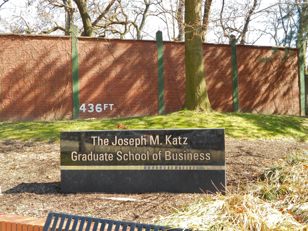Das Joseph M. Katz Graduate School of Business Gebäude mit einer Namensplakette im Vordergrund, einer Bank unten, Gras am Boden, einer Wand im Hintergrund, Bäumen und einem klaren blauen Himmel.