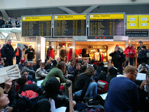 Eine große Gruppe von Menschen versammelt sich in einem Flughafen, einige sitzen mit Taschen und Papieren, andere stehen, mit Texttafeln, Schaufensterpuppen in Kleidern und Deckenlampen im Hintergrund, was auf eine Protestaktion hindeutet.