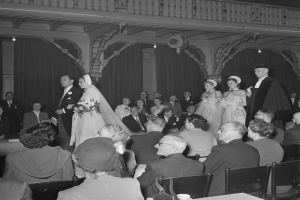 Schwarzes und wei├čes Foto einer Hochzeitszeremonie mit G├Ąsten, die auf Stühlen sitzen und stehen, Str├Ąu├čer und Gegenst├Ąnde auf Tischen, Vorh├Ąnge und Deckenleuchten im Hintergrund.