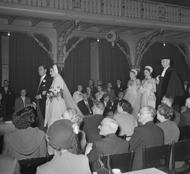 Schwarzes und wei├čes Foto einer Hochzeitszeremonie mit G├Ąsten, die auf Stühlen sitzen und stehen, Str├Ąu├čer und Gegenst├Ąnde auf Tischen, Vorh├Ąnge und Deckenleuchten im Hintergrund.