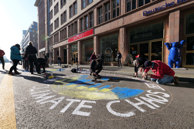 Eine Gruppe von Menschen sitzt vor einem Gebäude mit Fenstern und Namensschildern auf dem Boden, umgeben von Flaschen und anderen Gegenständen, mit Bäumen und einem klaren blauen Himmel im Hintergrund, während sie an einer Klimawandel-Demonstration in Berlin teilnehmen.