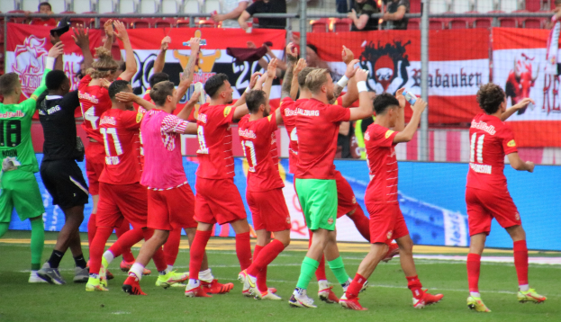 Gruppe von Fußballspielern, die ein Tor feiern, einige halten Flaschen und andere klatschen, mit einem Zaun, Bannern, Metallstangen, Stühlen und einem Dach mit Lampen im Hintergrund.