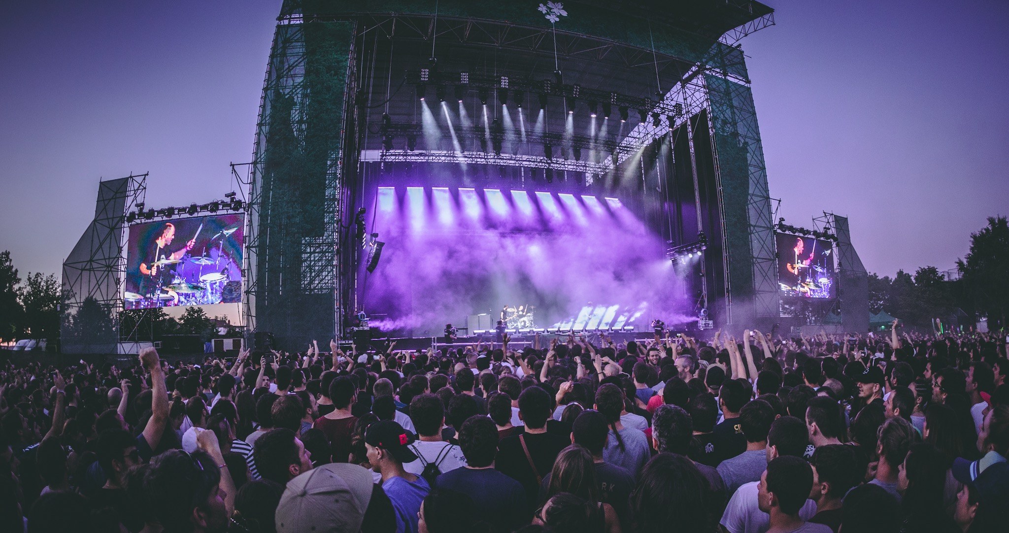 Eine große Menschenmenge auf einem Musikfestival mit einer Bühne, auf der Musiker spielen, beleuchtet von Lichtern und umgeben von Bildschirmen, vor einer Kulisse aus einem Gebäude, Bäumen und einem klaren blauen Himmel.