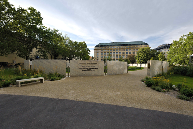 Eine Parkbank umgeben von Bäumen und Pflanzen, mit Gebäuden und einem bewölkten Himmel im Hintergrund und Text an der Wand hinter der Bank, wahrscheinlich in Zusammenhang mit dem Holocaust-Mahnmal in Berlin.