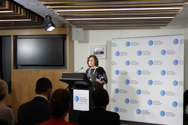 Eine Frau steht auf einer Bühne und spricht in ein Mikrofon, vor ihr sitzen Zuschauer, hinter ihr eine Wand mit einem Banner und einem Fernseher, an der Decke Lampen.