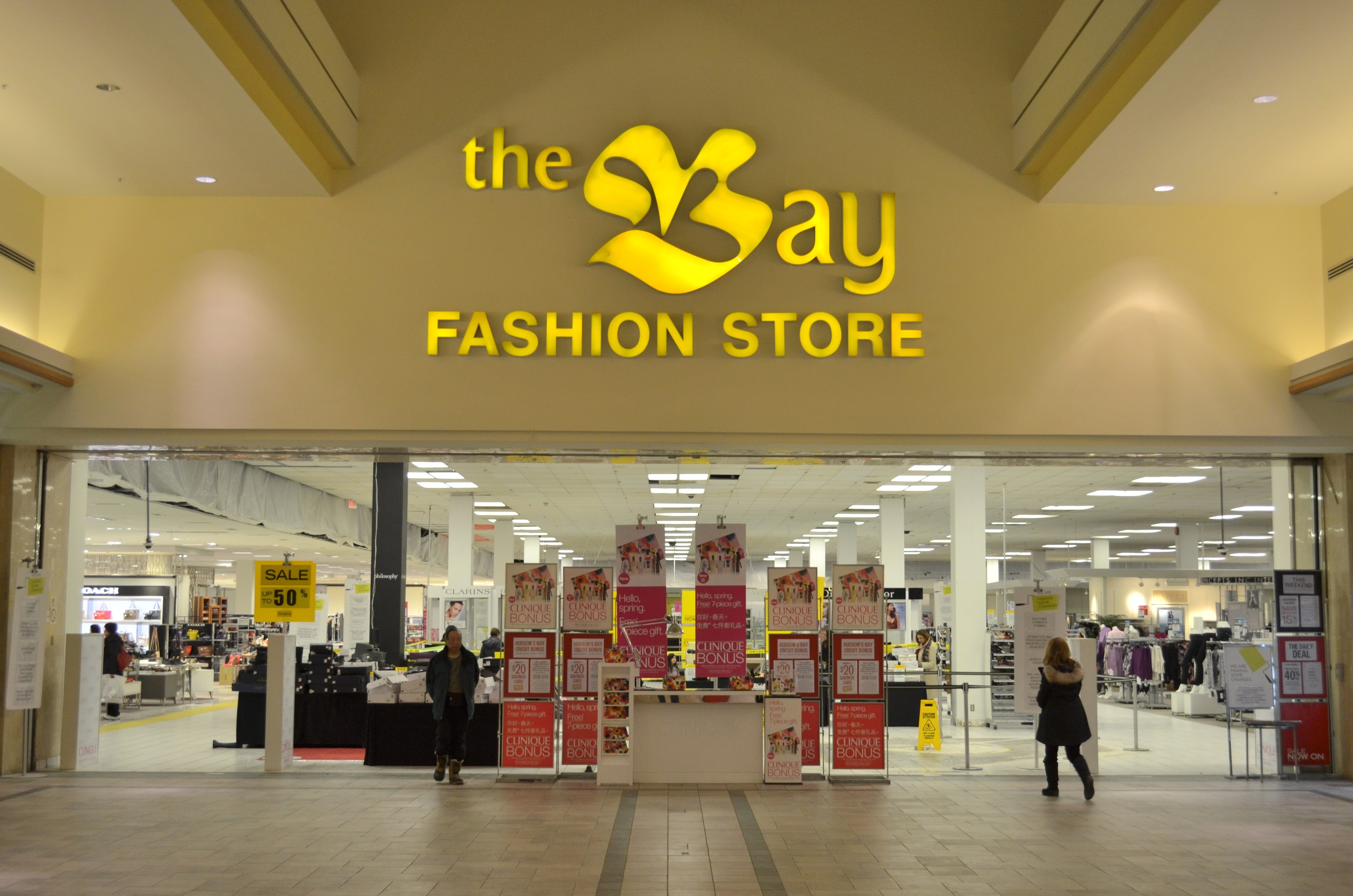 Außenansicht eines Bay Fashion Stores in einem Einkaufszentrum mit vorbeigehenden Kunden, Wandtafeln mit Text, Deckenleuchten und Hintergrundauslagen.