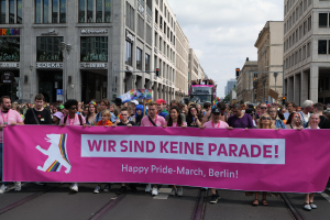 Eine Gruppe von Menschen marschiert auf einer Berliner Straße und hält eine pinkfarbene Fahne mit der Aufschrift "Happy Pride March", während Gebäude, Laternenpfähle und Verkehrszeichen die Straße säumen und der Himmel bewölkt ist.