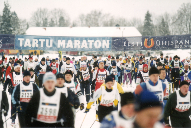 Gruppe von Menschen, die auf Skiern in einer schneebedeckten Landschaft mit Bannern, Bäumen und einem klaren blauen Himmel rennt, Text unten.