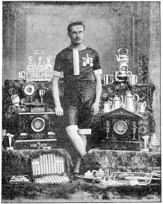 Ein Mann in einem Fußballtrikot steht stolz vor einem Tisch voller Trophäen und Pokale, mit der Aufschrift "1909-1910 Weltmeisterschaftssieger" am unteren Rand des Schwarz-Weiß-Fotos.