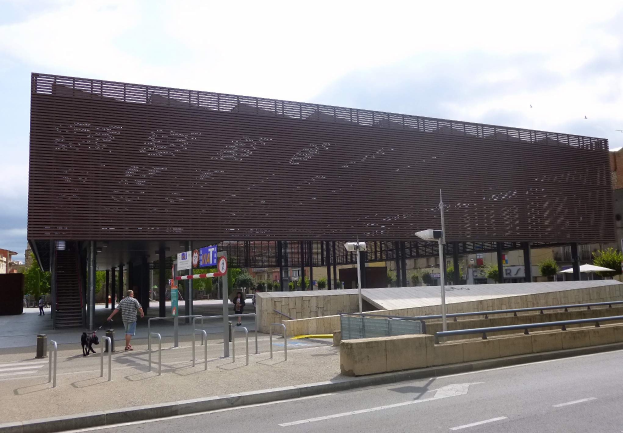 Großes städtisches Einganggebäude mit vielen Fenstern, umgeben von Straßeninfrastruktur, Fahrzeugen, Fußgängern, Bäumen und einem bewölkten Himmel.