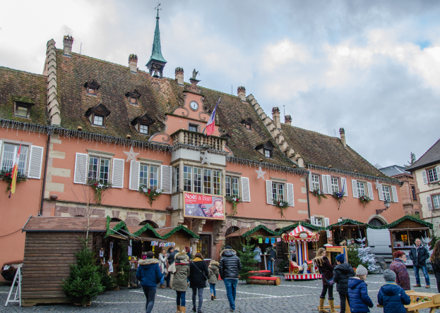 Eine Gruppe von Menschen geht eine Kopfsteinpflasterstraße in einem kleinen Dorf entlang, umgeben von Gebäuden mit Fenstern, Pflanzen, Flaggen und einer Tafel mit Text, unter einem bewölkten Himmel, einige tragen Taschen.