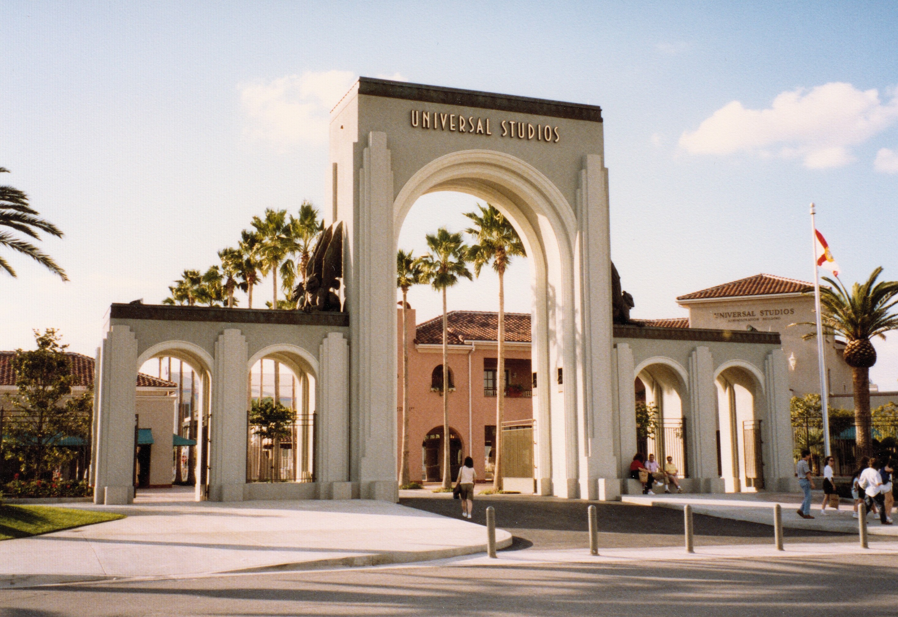 Eingang zu Universal Studios Hollywood mit einem Bogen mit Text, einer Statue, Menschen, Bäumen, Gebäuden, einem Fahnenmast mit einer Fahne und einem bewölkten Himmel.