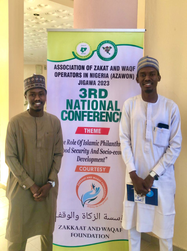 Zwei Männer vor einem Banner mit der Aufschrift "3. Nationalkonferenz" mit einer Säule und Deckenleuchten im Hintergrund, wahrscheinlich an der Veranstaltung im Jahr 2023 teilnehmend.