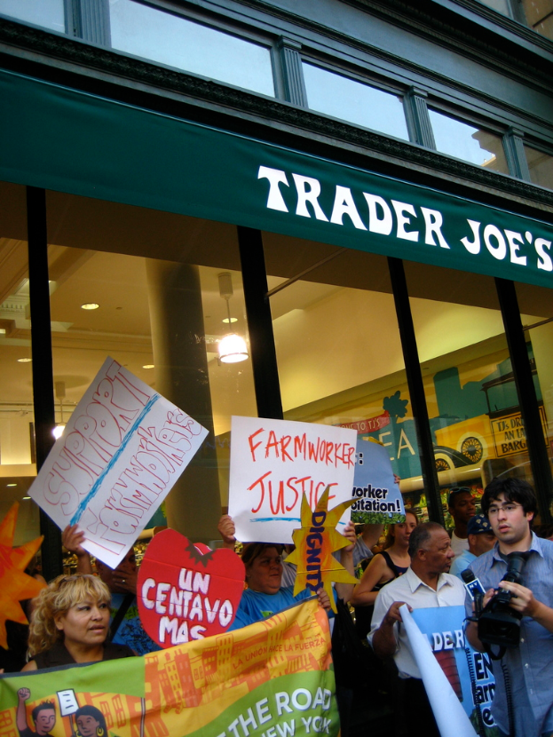Eine Gruppe von Menschen steht vor einem Geschäft und hält Schilder und Plakate hoch, wobei hinter ihnen eine Säule und Lichter auf dem Dach zu sehen sind.