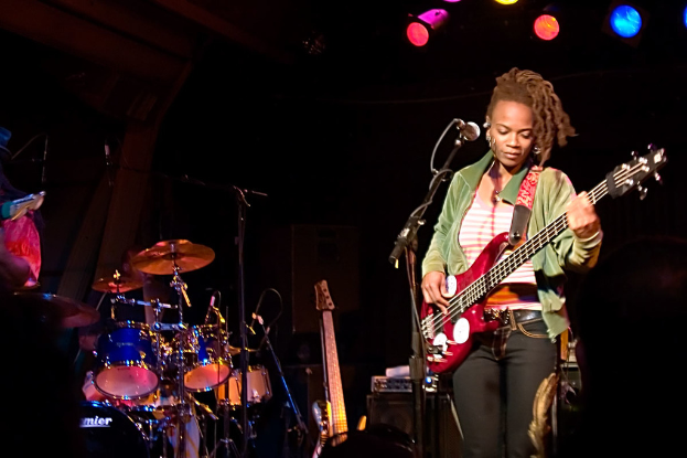 Eine Frau in einer grünen Jacke spielt Gitarre auf der Bühne während eines Konzerts, mit einer Bandaufstellung zu ihrer Linken und Lichtern am oberen Ende des dunklen Hintergrunds.