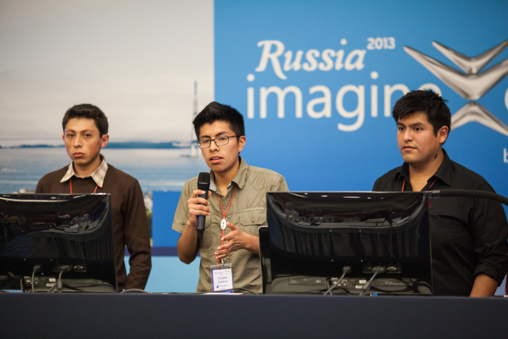 Drei Jungen stehen neben einem Schreibtisch mit zwei Computersystemen, einer spricht in ein Mikrofon, trägt ID-Karten mit roten Tags, vor einer Wand mit einem Werbeplakat, auf dem 'Russia Imagine 2013' steht.