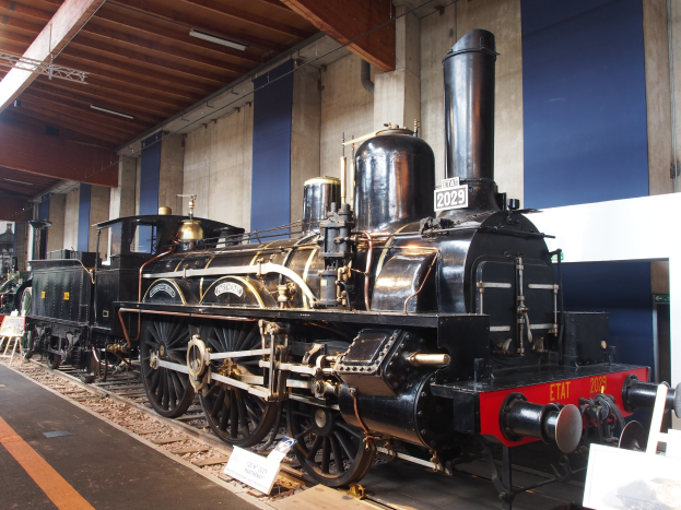Eine gro├če, alte Dampflokomotive in einem Museums Setting umgeben von Eisenbahnschienen, Informationsschildern und anderen Ausstellungen, mit einer Wand und Deckenbeleuchtung im Hintergrund.