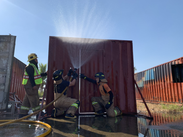 Feuerwehrleute in Helmen und Handschuhen sprühen Wasser aus einem Schlauch auf einen brennenden Metallbehälter, mit Bäumen und einem klaren blauen Himmel im Hintergrund.