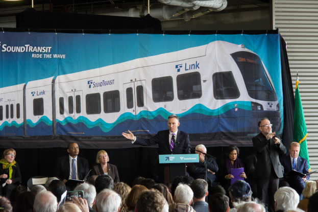 Ein Mann in Anzug und Krawatte spricht in ein Mikrofon an einem Rednerpult, mit einem Banner mit einem Zugbild und Text dahinter, einem Verschluss und einer Flagge rechts und Lampen und Rohren an der Decke.
