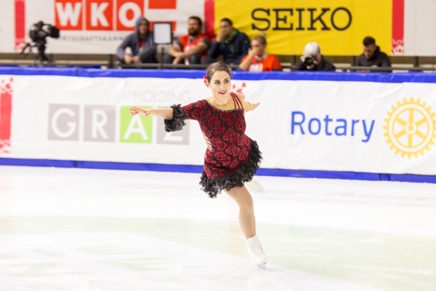 Eine Frau in einem roten und schwarzen Kleid gleitet elegant über das Eis während der Weltmeisterschaften im Eiskunstlauf. Im Hintergrund sind Zuschauer und ein Kameramann zu sehen.