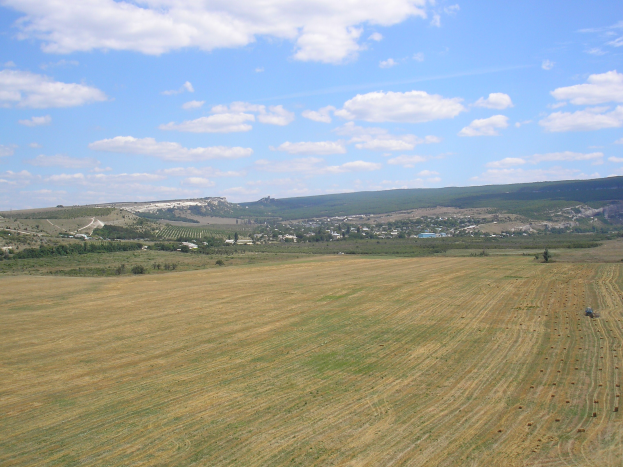 Ein Traktor, der auf einem bewirtschafteten Feld umgeben von Bäumen, Gebäuden, Hügeln und einer wolkenverhangenen Himmel operiert.