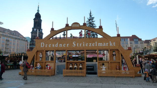 Weihnachtsmarkt in Dresden, Deutschland mit Menschen, die spazieren gehen, einem geschmückten Bogen, Bäumen, Gebäuden und bewölktem Himmel.