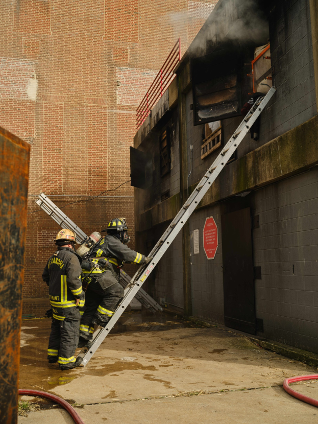 Feuerwehrleute mit Helmen klettern eine Leiter hinauf zu einem raucherfüllten Gebäude, mit einem Metallobjekt links und Rohren an der Basis.