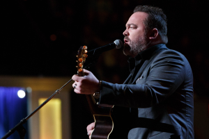 Ein Mann in einem Anzug spielt Gitarre und singt in ein Mikrofon auf einer schwach beleuchteten Bühne, mit Dierks Bentley im Hintergrund.