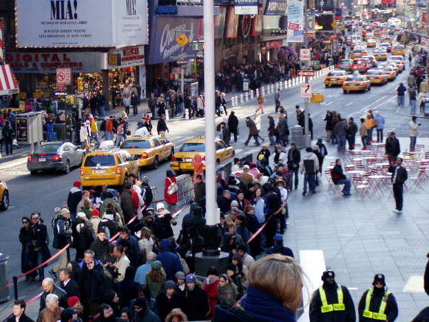 Fahrzeuge passieren eine Straße mit Fußgängern auf den Gehwegen, umgeben von Stühlen, Tischen, Laternen, Schildern und Ampeln, mit Gebäuden und Geschäften auf beiden Seiten.