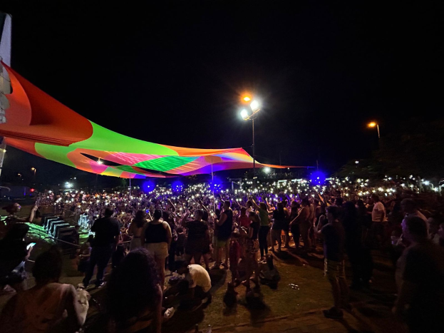 Eine große Menschenmenge steht vor einem hell erleuchteten Zelt in der Nacht während des 2018er Kunstfestivals, mit bunten Lichtern und Pfählen, die die Szene beleuchten, einem Gebäude auf der linken Seite und dem Nachthimmel im Hintergrund.