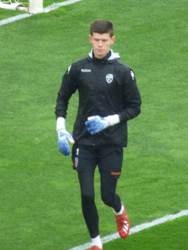 Ein Mann in einer schwarzen Jacke und blauen Handschuhen läuft auf einem Fußballfeld, im Hintergrund ist ein Tor zu sehen.