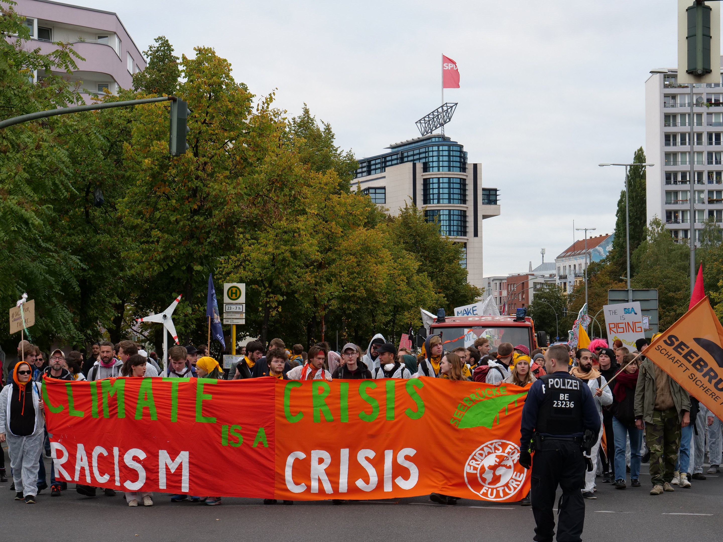 Eine Gruppe von Menschen marschiert mit einem "Klimakrise ist eine Krise"-Schild die Straße entlang, mit Bäumen, Laternen und parkenden Fahrzeugen am Straßenrand, Gebäude im Hintergrund unter einem klaren blauen Himmel.